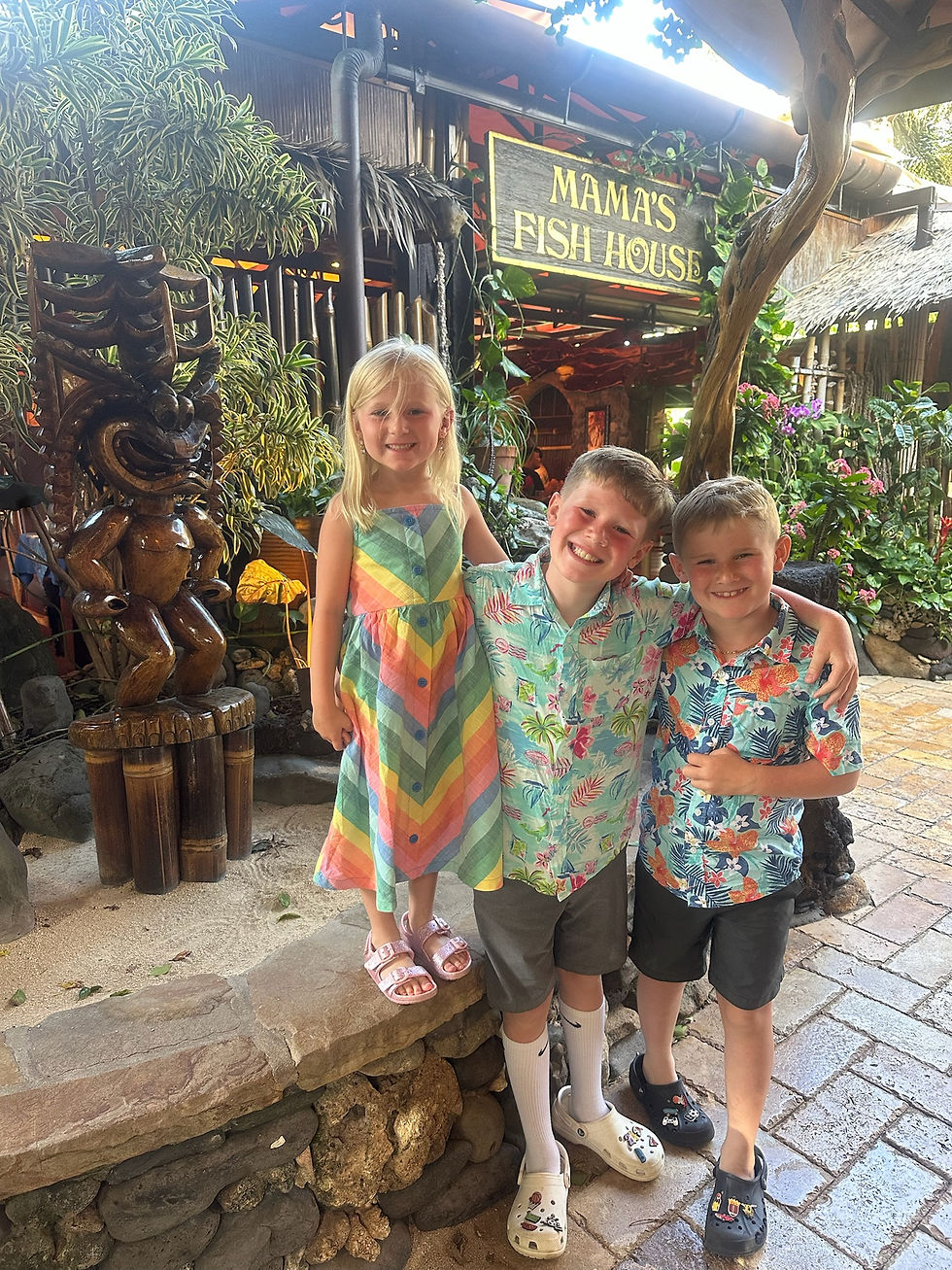 Three smiling children in colorful outfits stand by a wooden statue at Mama’s Fish House, surrounded by lush greenery and vibrant flowers.