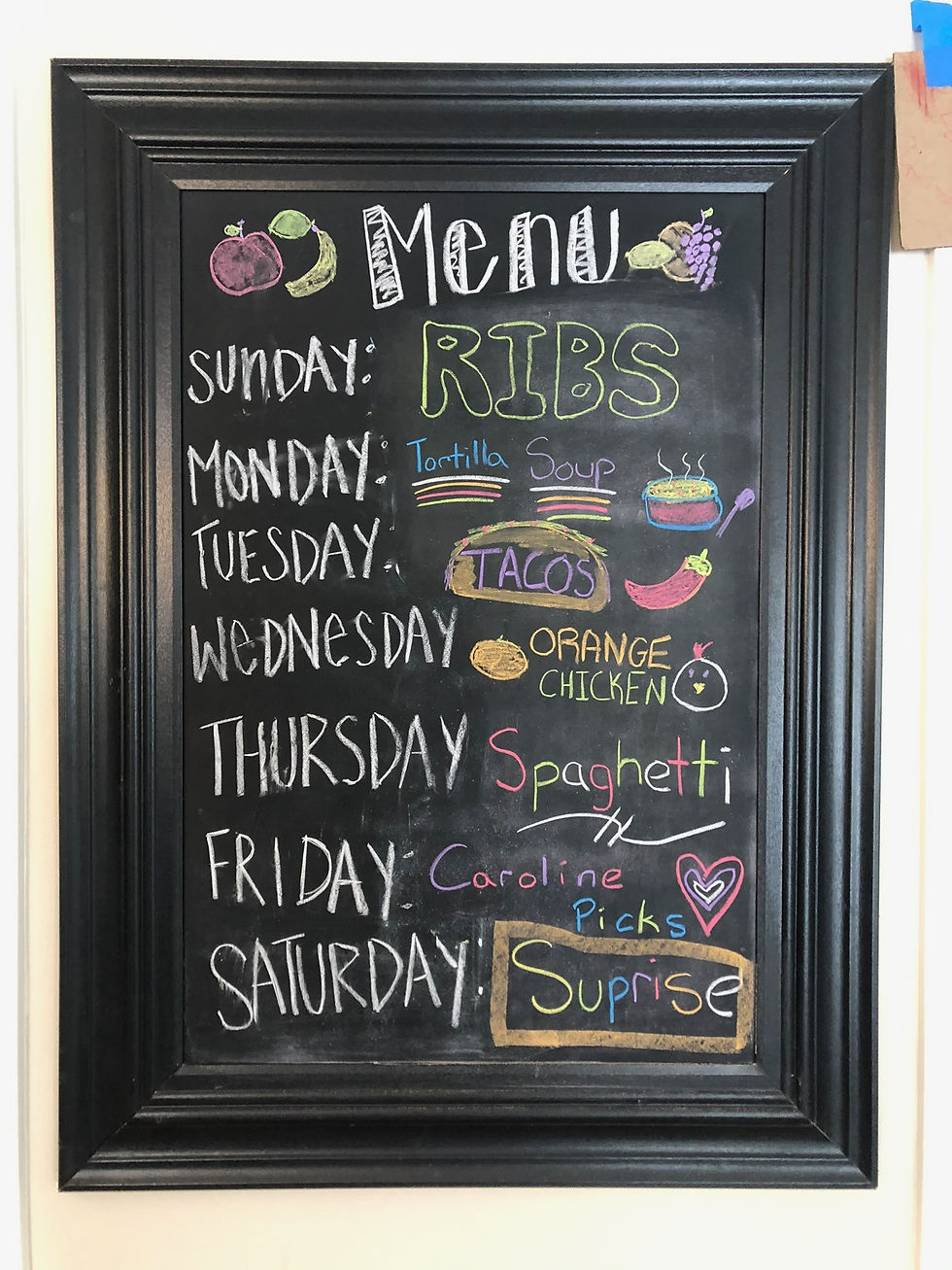 Chalkboard menu with weekly meals listed: ribs, tortilla soup, tacos, orange chicken, spaghetti, "Caroline Picks," and "Suprise." Colorful doodles.