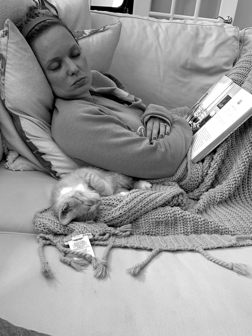 Woman sleeping on a couch with a book and a cat. Wrapped in a knitted blanket in a cozy, serene setting. Monochrome image.