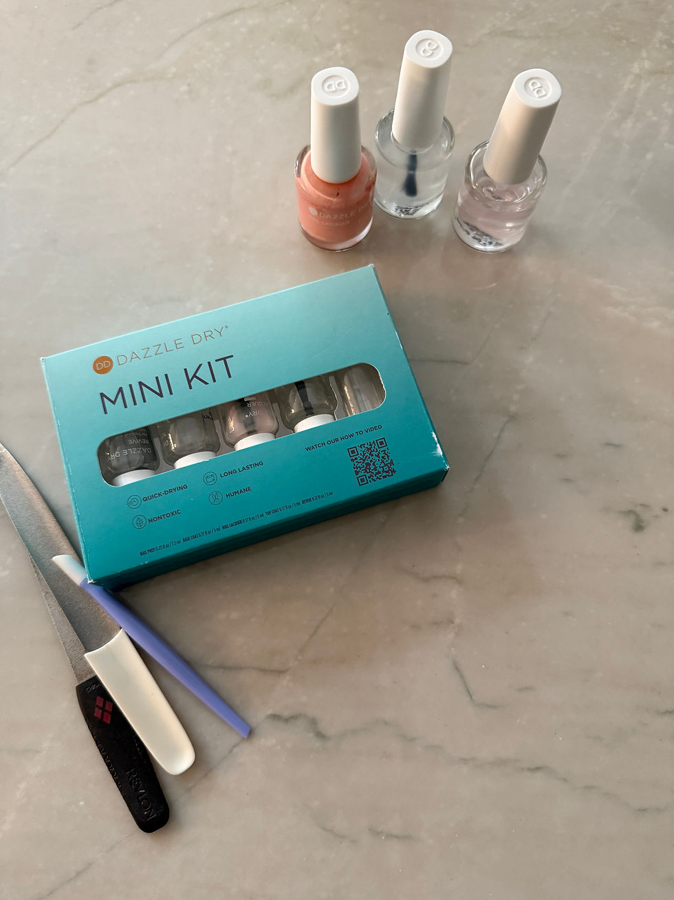 Blue "Dazzle Dry Mini Kit" box with three nail polish bottles, a nail file, and tools on a marble surface.