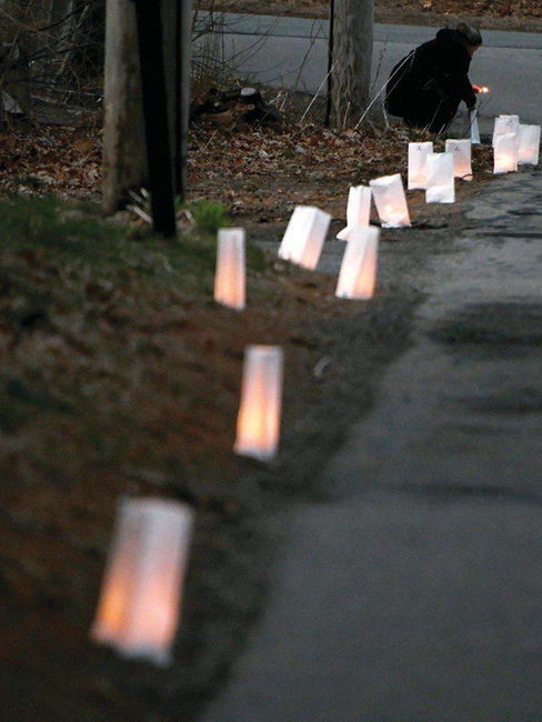 Lit paper lanterns line a dim path at dusk, casting a warm glow. A person kneels in the background, lighting another lantern.