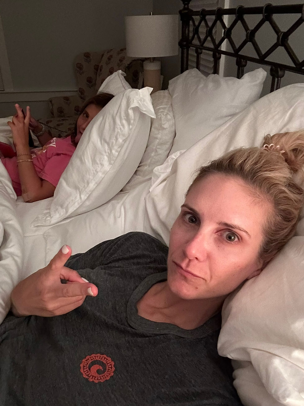 Two people lie in bed; one takes a selfie while the other looks at a phone. Calm mood, white bedding, dimly lit room.