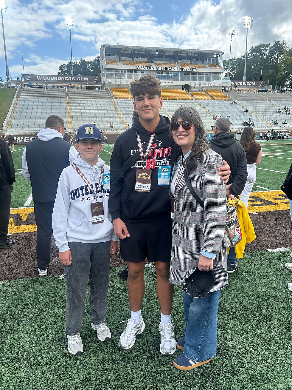 Three people pose on a football field at Western Michigan University. A boy in a cap wears a white sweater, while the others wear dark tops.