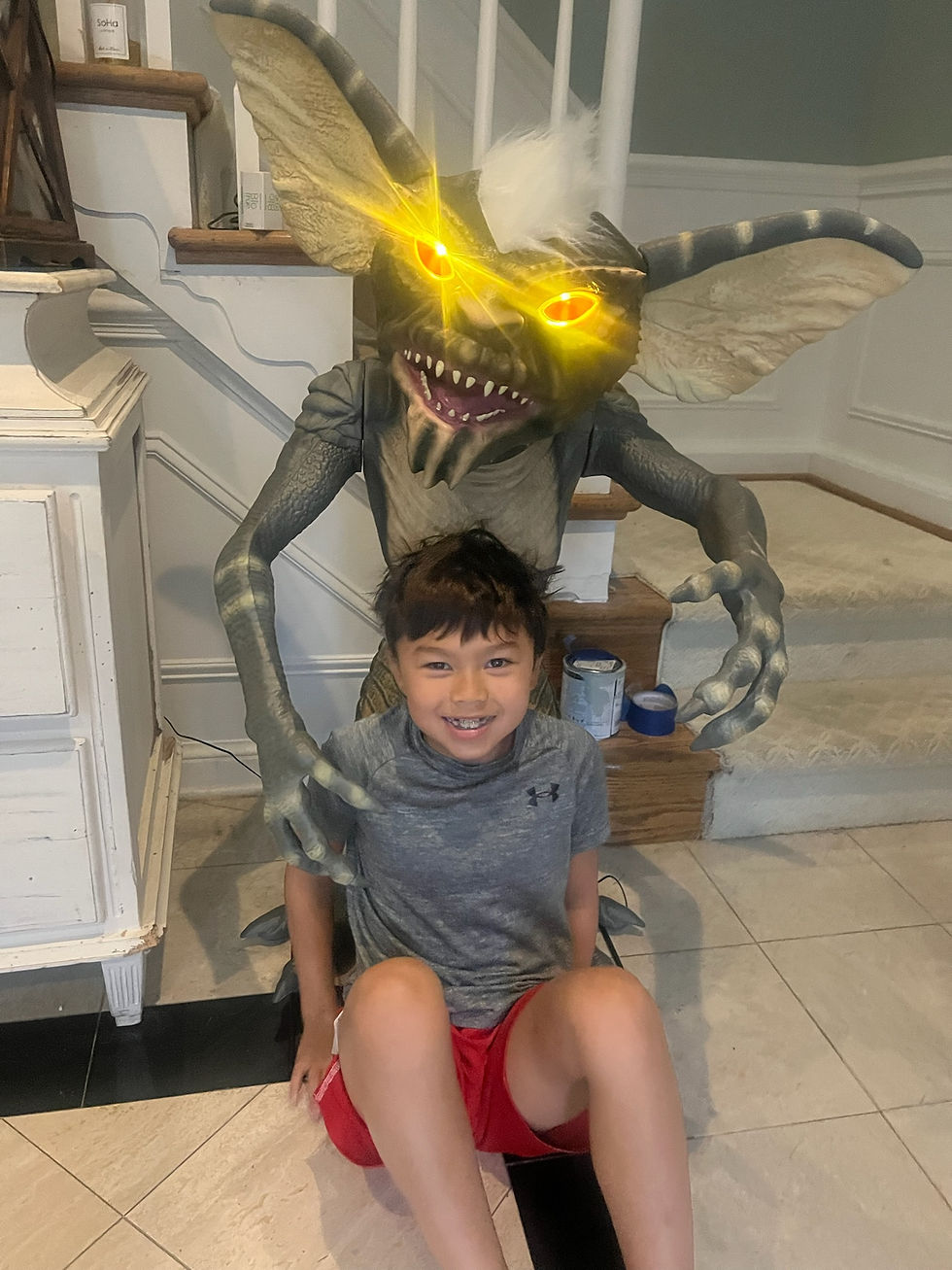Child in gray shirt and red shorts smiles while sitting in front of a gremlin figure with glowing eyes. Indoors by a staircase.