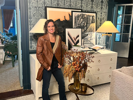 Smiling woman in a brown leather jacket stands in a stylish living room with framed art, lamps, and patterned wallpaper.