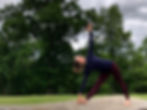 Woman in a navy top and maroon leggings performs yoga outdoors on a rock, surrounded by lush green trees under a cloudy sky.