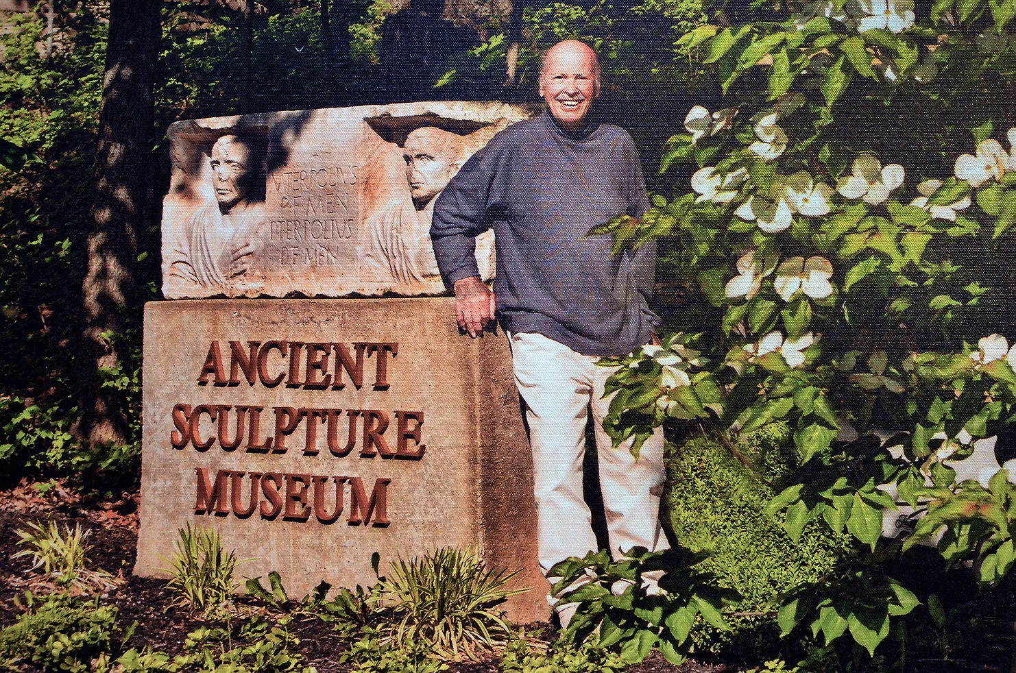Man stands smiling and leaning against a large sign that reads 'ancient sculpture museum'