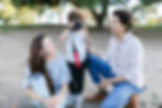 Child with curly hair takes a photo with a camera, flanked by smiling adults in casual clothes. Sunny park setting with trees in the background.