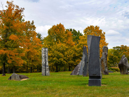 Stone sculptures stand on a grassy field with trees in autumn colors. The sky is cloudy, creating a calm and serene atmosphere.