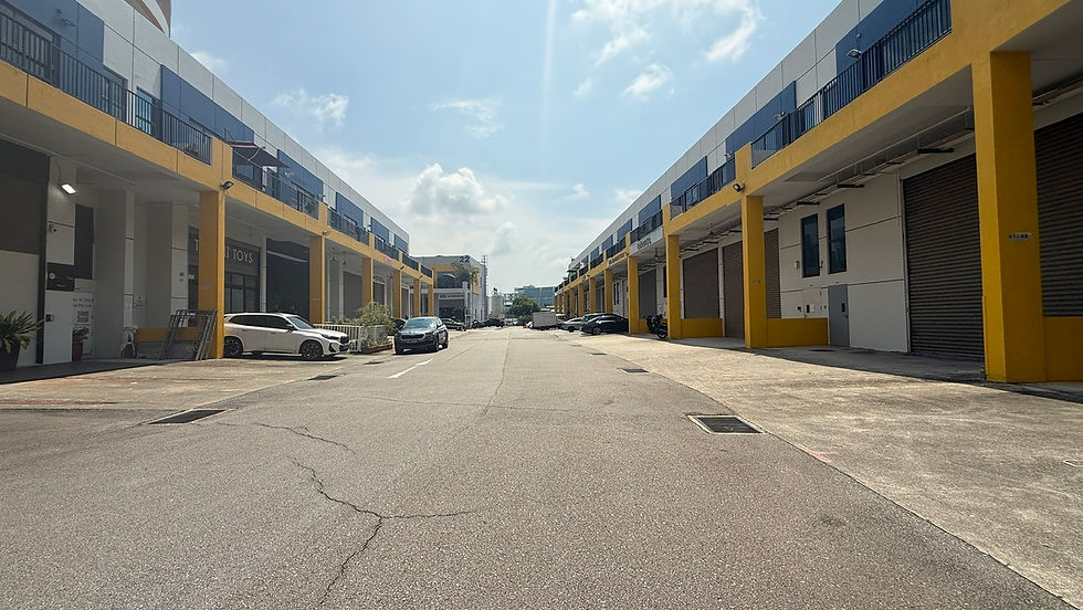 Industrial area with yellow buildings and parked cars under a clear sky. Buildings have roll-up doors; a few clouds are visible. Quiet atmosphere.