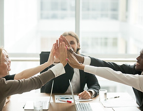 A diverse group of business professionals in a modern office, enthusiastically joining hands for a group high-five, symbolizi