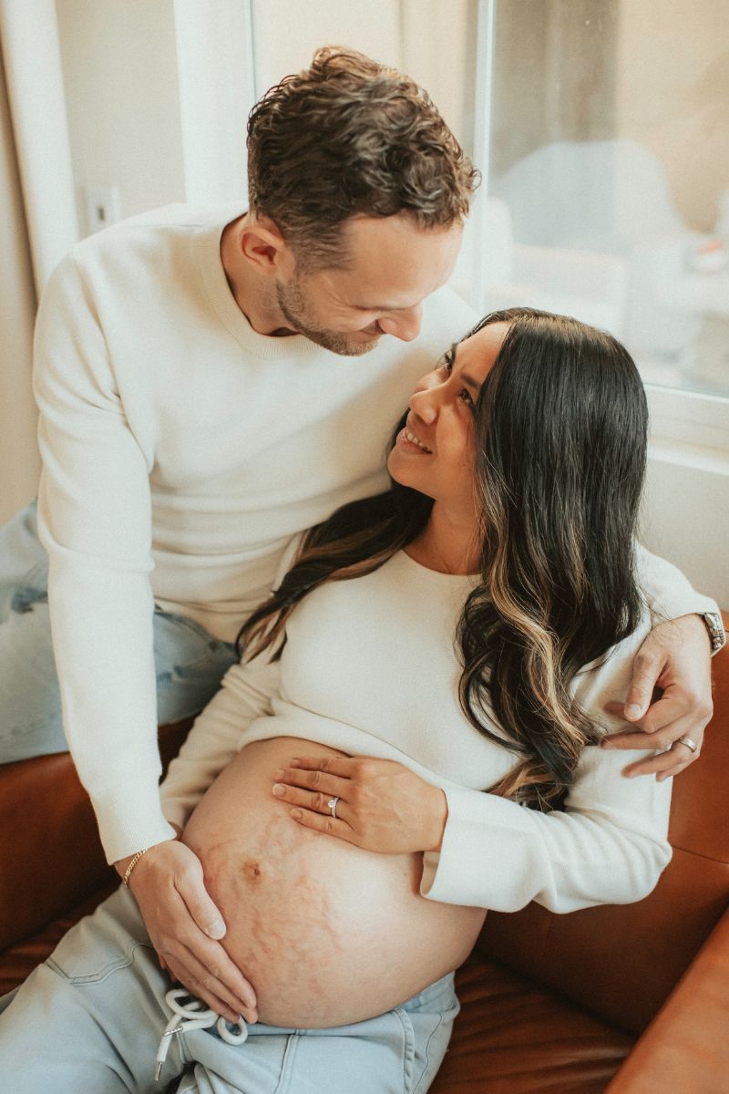 Future mom and dad snuggling on the couch wearing jeans and white tops