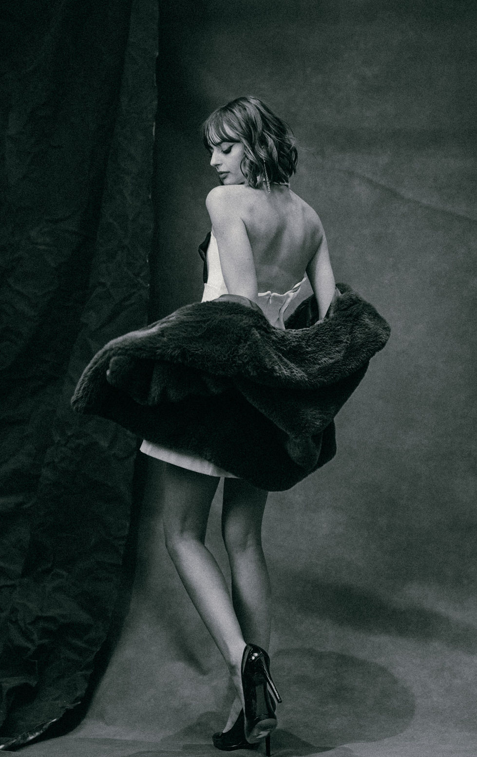Black-and-white photo of a woman viewed from behind, in a low-draped fur coat over a white skirt and heels.