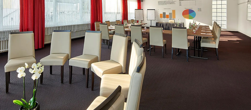 Konferenzraum mit U-förmiger Tischaufstellung und beigen Stühlen, Sonnenlicht fällt durch rote Vorhänge.