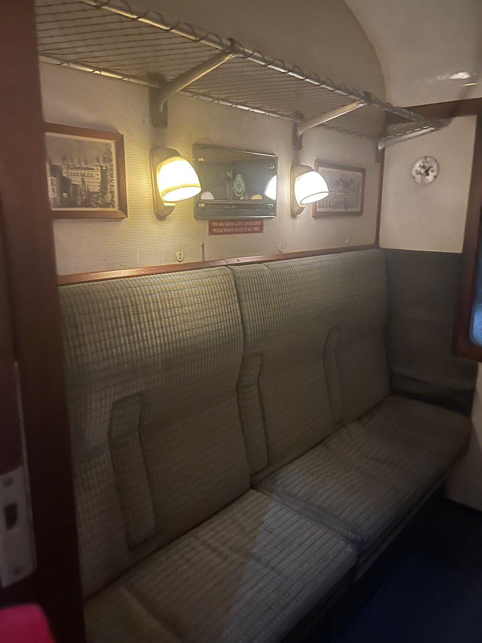 Inside shot of cubicle on a train featuring a bench with two lamps above and a large rack for suitcases across the top. There is a window to look outside as well.