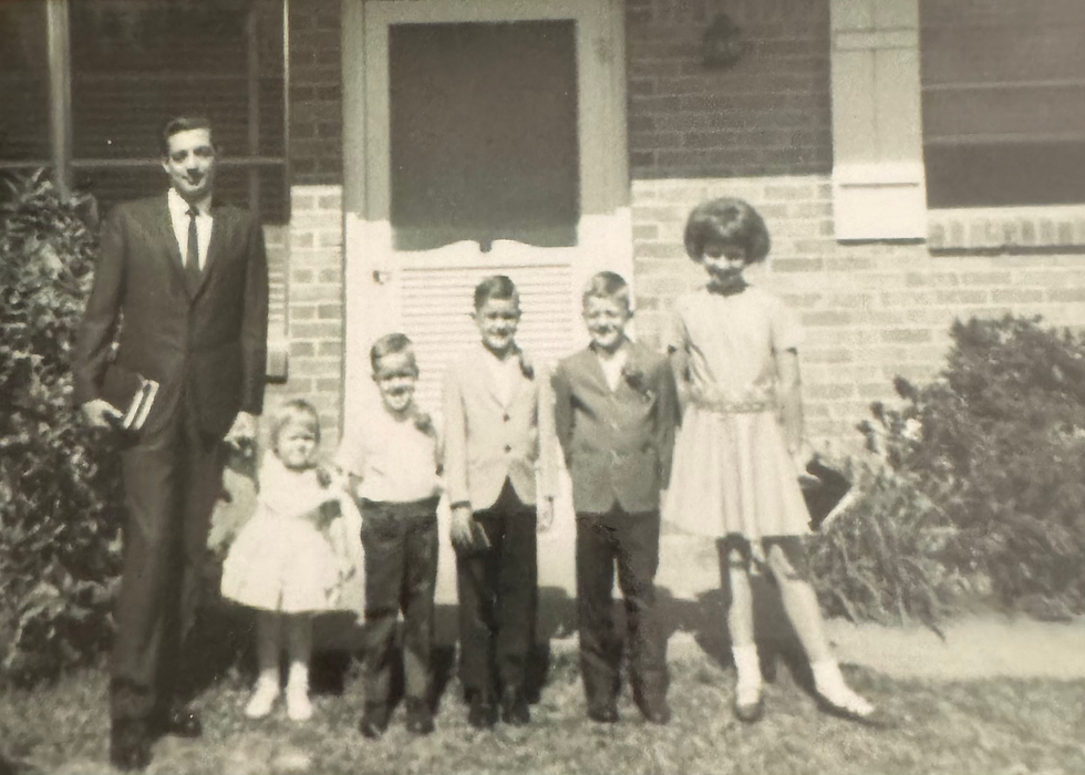 A family is posed for a picture in front of small house. There is a man with 2 girls and three boys all dressed up for church.