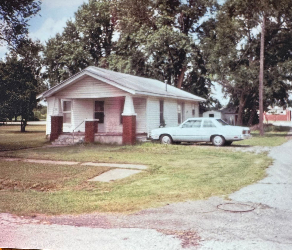 An older house from the 50's with a car parked outside. The house has a large front porch and a small yard with a sidewalk.