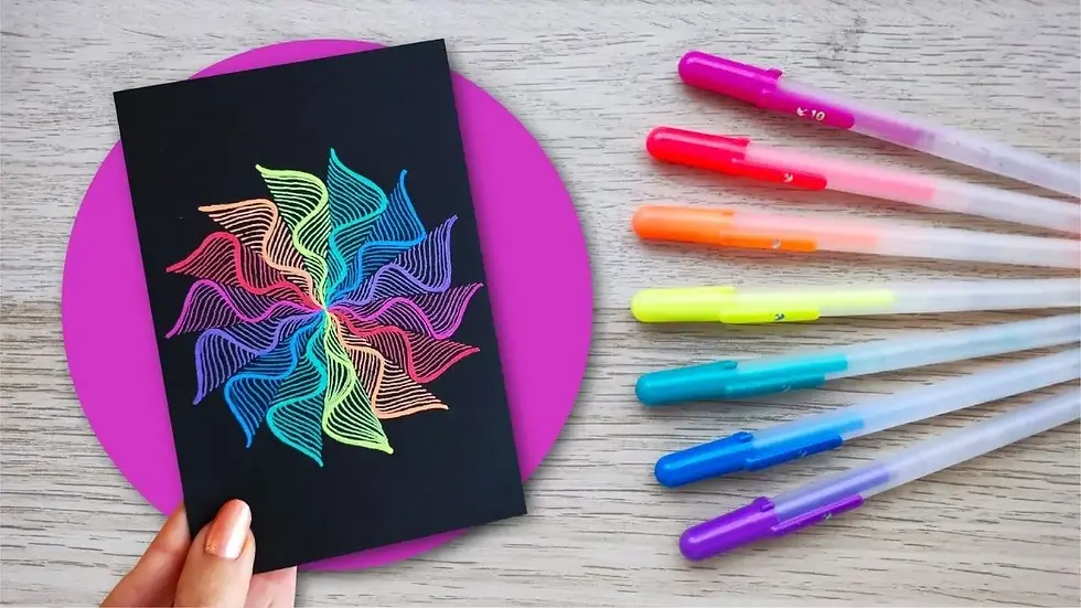 Mano sostiene una cartulina negra con un mandala espiral de colores neón; a la derecha, seis bolígrafos de gel de colores sobre una mesa de madera clara.