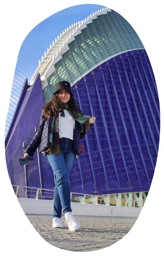 Ro sonriente de pie al aire libre frente al gran edificio moderno Hemisféric de Valencia con amplias fachadas de vidrio bajo cielo azul