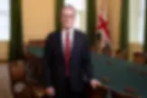 Un hombre de cabello canoso y gafas está de pie junto a una mesa de reuniones, con una bandera británica detrás.