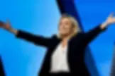 Mujer rubia de mediana edad sonríe con los brazos extendidos, lleva chaqueta oscura y está frente a un fondo azul brillante.