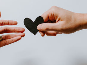 A hand holding a black paper heart reaching toward an open hand.
