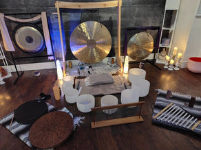 Meditation room with gongs, crystal bowls, and candles on wooden floor. Soft lighting creates a serene atmosphere. Instruments on striped mats.
