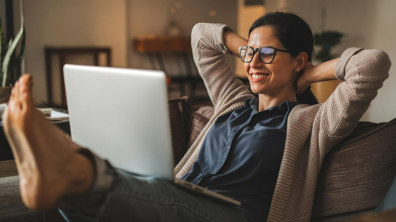 They color photograph of a woman leaning with her arms behind her head and a laptop in her lap looking relaxed