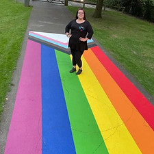 A picture of Michele, a Trustee of CrawleyLGBT. Michele is stood on a large painted rainbow progress flag. She is wearing a black CrawleyLGBT top.