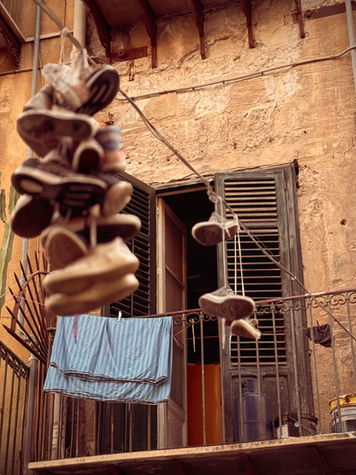 Fine art photograph of worn shoes hanging from a balcony against a weathered Italian façade in warm, earthy light.