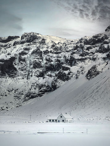 Photograph of a small farmhouse surrounded by snow beneath rugged Icelandic cliffs.