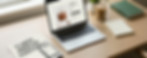 Wide lifestyle scene on a light wood desk near a window. A laptop open to a clean Shopify product page.