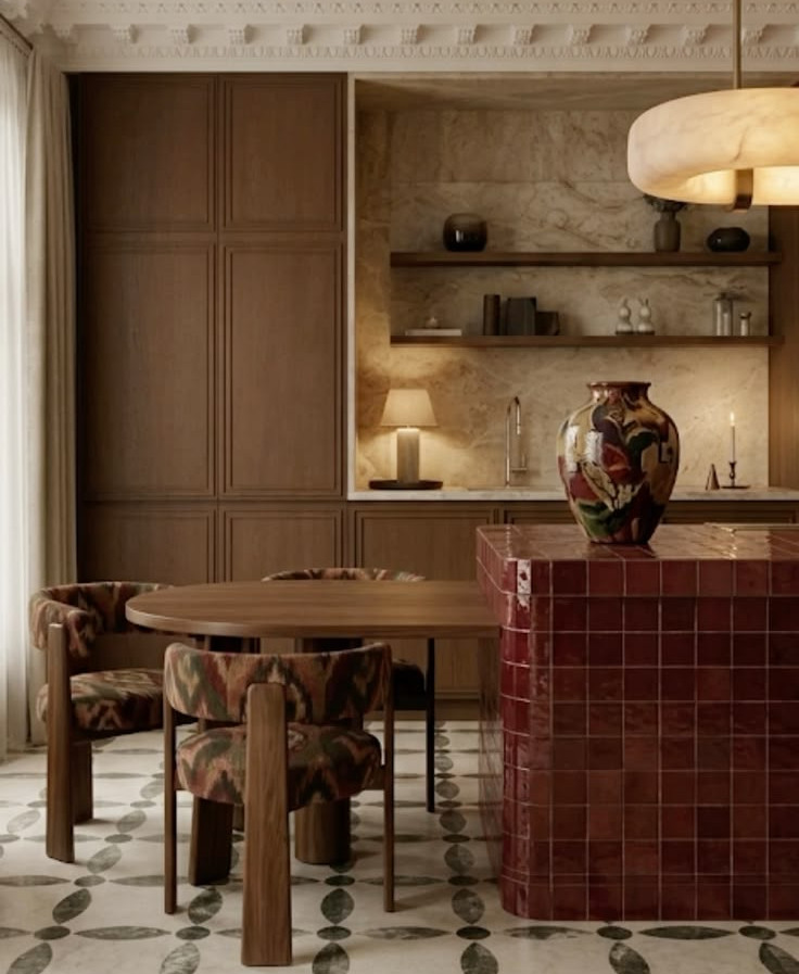 Warm modern dining room with round wood table, patterned chairs, red tiled island, shelves, lamp, and large vase.
