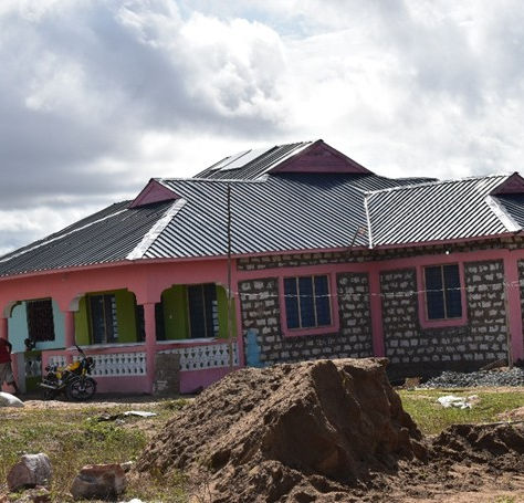 Colorful single-story house with pink trim and black roof in a rural setting. A person and a motorcycle are in front. Cloudy sky above.