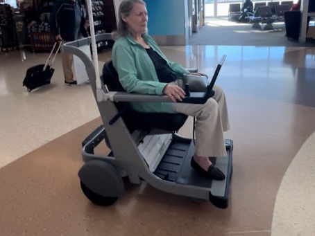 An older woman seated in an autonomous mobility vehicle inside an airport terminal, moving independently along the concourse. Other travelers and seating areas are visible in the background.