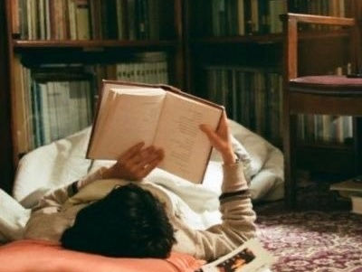 Person lying on a bed reading an open book in a cozy book-lined room with a patterned rug.