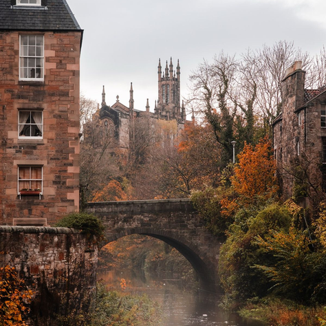 Stone bridge over misty river between old houses, with autumn trees and a Gothic church tower under gray sky.