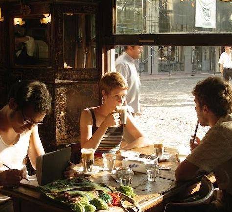 Three people sit at a sunlit cafe table; one uses a laptop, one sips a drink, and one smokes near food and glasses.