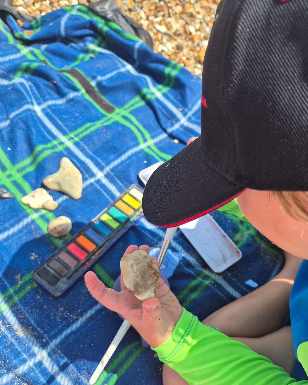 My child painting a pebble with watercolours while sitting on our beach blanket