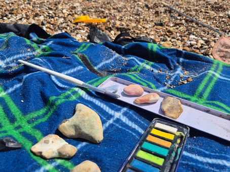 Watercolour paints, brushes and painted pebbles on a blue beach towel beside the sea