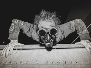 Mężczyzna w demonicznej masce i goglach pochyla się nad parapetem, wystawiając język.