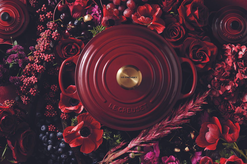 Panela Le Creuset vermelha, pegador dourado, entre flores e frutas escuras.