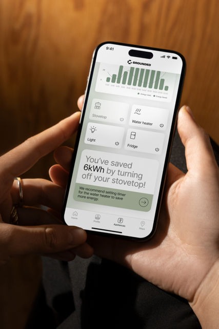 Photo looking over a woman's shoulder at her iPhone, showing a Grounded+ app screen revealing information on her energy usage. The main focus on the screen is a widget telling her how much power she saved by turning off her stovetop. 
