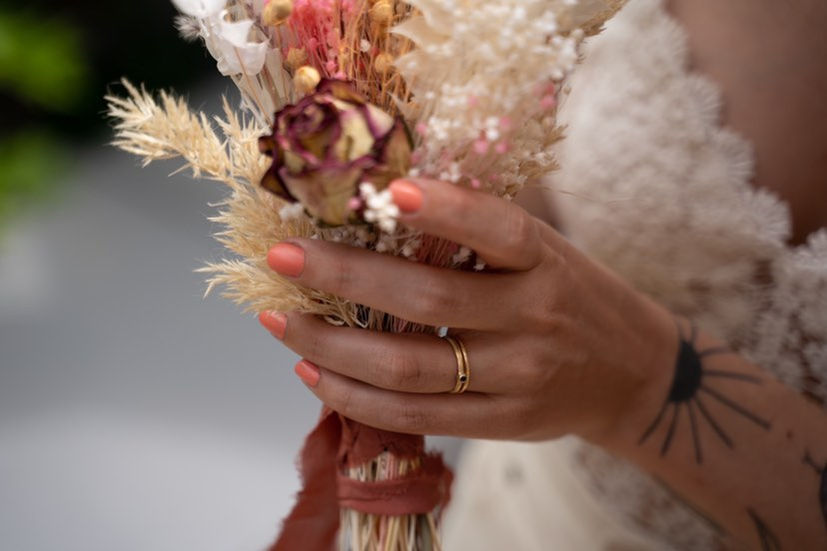 Close-up van de hand van een bruid, met in haar hand een boeket droogbloemen van Dryflowerbar en aan haar vinger een geelgouden trouwring, gemaakt door Atelier VogelVrij.