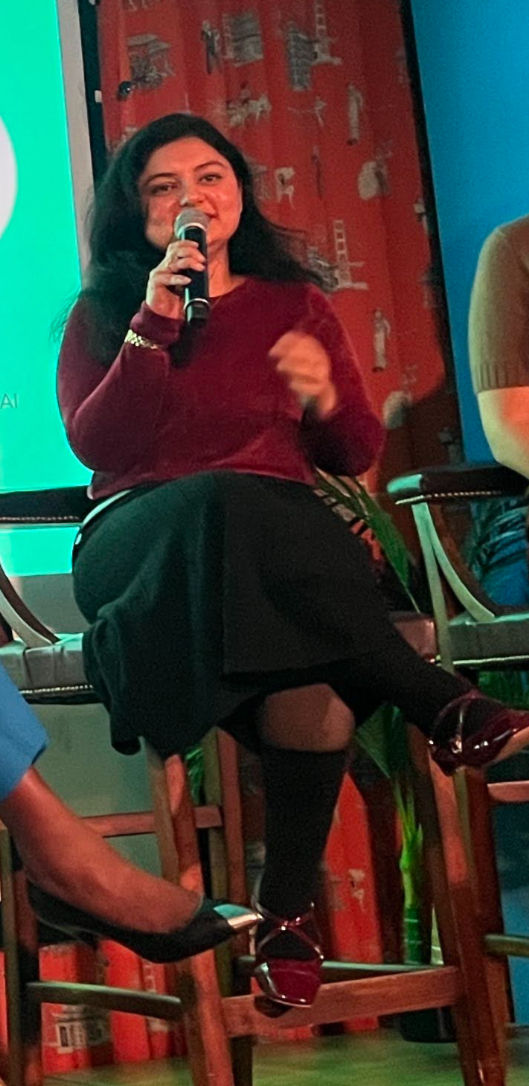 Woman in a maroon top speaks into a mic while seated on a panel. Red patterned curtain backdrop. Bright, engaging mood.