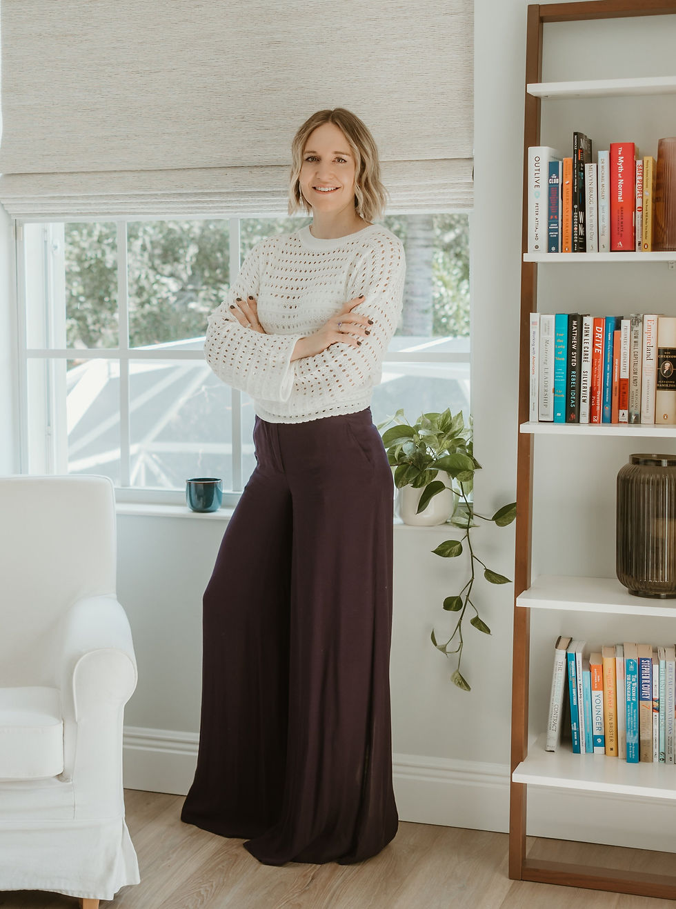 Smiling woman in white sweater and dark pants stands by a bookshelf with colorful books. Bright room with window and plant. Cozy vibe.