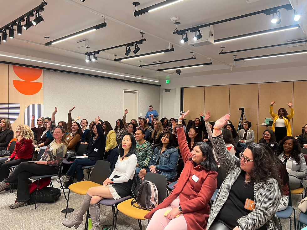 Audience of mostly women raise hands in a bright modern conference room during an upbeat meeting.