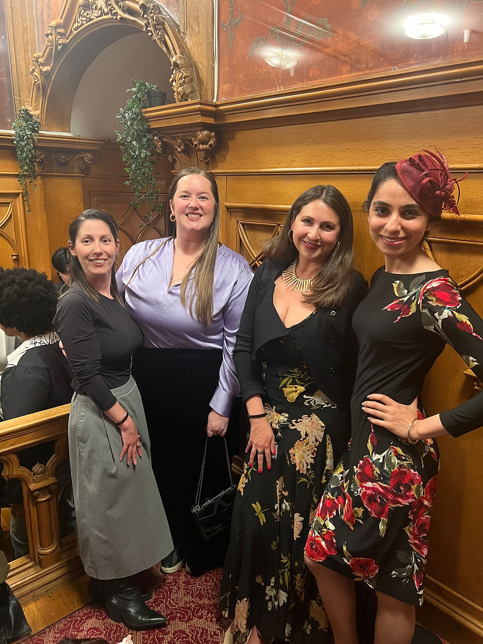 Four women pose and smile in an ornate wooden interior. One wears a red floral dress and hat. The setting is elegant and warm.