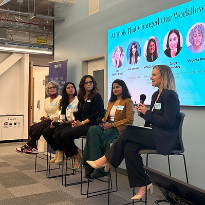Five women sit on a panel discussing AI tools in an office setting, with a screen displaying their names and roles. One speaks into a microphone.