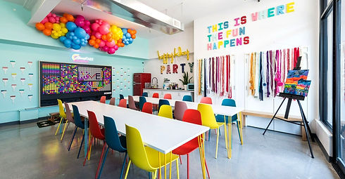 A colorful room with balloons on the ceiling and a sign 'this is where the fun happens', plus 2 long white tables with different colored plastic chairs